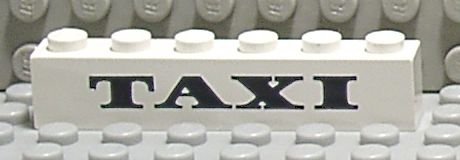 White LEGO 3009pb002 1x6 brick with black 'TAXI' thick pattern, on a gray baseplate.