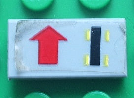 White LEGO 1x2 tile with sticker showing a red straight arrow on the left and a black car chassis symbol with yellow wheels on the right.
