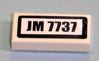 White LEGO 1x2 tile with 'JM 7737' pattern on a black-bordered sticker.