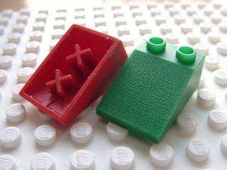Red LEGO Minitalia Slope 33 3x2 brick showing bottom X supports, next to a green Slope 33 3x2 brick on a white LEGO baseplate.
