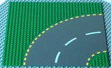 Green LEGO 32x32 road baseplate with dark gray curved road, white dashed lane lines, and yellow dashed edge lines.