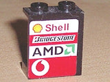 A black LEGO 1x2x2 panel with hollow studs, featuring a sticker with Shell, Bridgestone, AMD, and Vodafone logos.