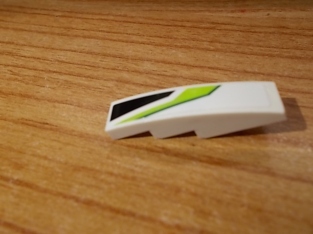 Close-up of a white LEGO curved slope 4x1, featuring a green and black diagonal pattern on its right side, on wood.