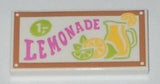 White LEGO 2x4 tile with a brown border and 'LEMONADE' pattern. Features pink text, '1,-' price, a lemonade pitcher, and citrus slices.