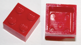 A shiny red LEGO 2x2 brick shown from two angles: one displaying its four studs on top, and another revealing its hollow underside without tubes.