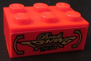 Close-up of an orange LEGO 2x3 brick featuring a black sticker with gold 'Brads TOWING 730-108' text.