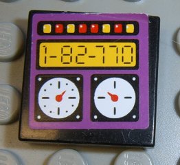 Close-up of a black LEGO 2x2 tile with a purple sticker showing a digital display '1-82-770', two gauges, and colored lights.
