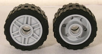 Two LEGO wheels, light gray rims with black tires. One shows spokes, the other shows fake bolts and a pin hole.